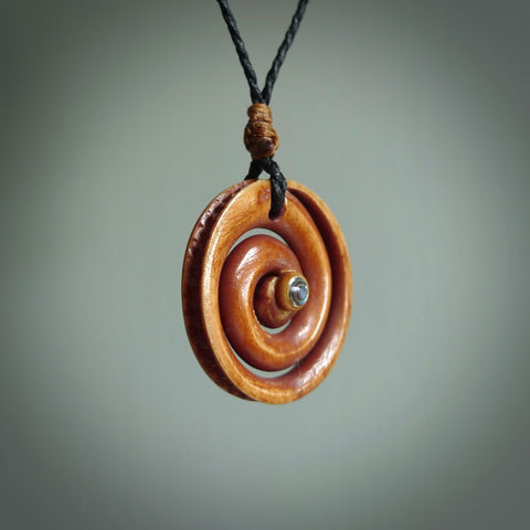 A hand carved and intricate koru pendant made for us by Yuri Terenyi. This is a beautiful little piece and is emblematic of the well known and loved Koru design. It is carved from stained bone in a hollowed, koru spiral shape with paua shell inlay in the middle of the koru. It is suspended from a black cord with a gingernut floret and the necklace is adjustable.