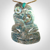 This photo shows a large hand carved Aotea stone Tiki pendant. It is a very light blue colour with pāua shell insert eyes. The cord is a thick camel colour and is length adjustable