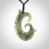 This picture shows a large hand-carved contemporary koru pendant. It is carved from New Zealand Kokopu jade and is suspended from a hand-plaited, black cord.