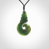 This picture shows a hook shaped pendant, hand carved from New Zealand flower jade. The top of the hook, the shaft, is a twist shape. We will provide this with an adjustable plaited cord. The cord is a adjustable and is a black colour. Twist and koru designs are also displayed in this stunning New Zealand Marsden Flower Jade pendant. Hand made by Ana Krakosky.