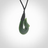 This photo shows a greenstone hook, or matau, pendant. It is a beautiful green British Columbia jade stone. The plaited cord is a black colour. It is plaited and the length can be adjusted.