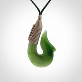 This photo shows a greenstone hook, or matau, pendant. It is a beautiful green British Columbia jade stone. The plaited cord is a green colour. It is plaited and the length can be adjusted.