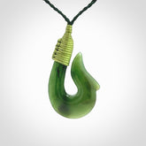 This photo shows a greenstone hook, or matau, pendant. It is a beautiful green British Columbia jade stone. The plaited cord is a green colour. It is plaited and the length can be adjusted.