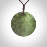 This piece is a large sized, oval round, disc pendant. It is suspended on a brown coloured braided cord that is length adjustable.