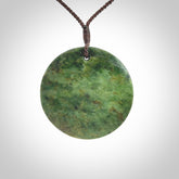 This piece is a large sized, oval round, disc pendant. It is suspended on a brown coloured braided cord that is length adjustable.