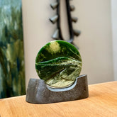 A spectacular, engraved jade disc sculpture. Hand carved in New Zealand from stunning Jade. This is a beautiful disc sculpture carved with a mountain and valley into its face. It is presented with a greywacke beach stone display stand. A beautiful sculptural piece hand carved in New Zealand.