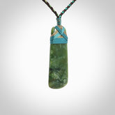 This photo shows a beautiful New Zealand jade toki suspended from a hand plaited adjustable cord. The binding around the top of the toki is a blue and bronze colour. The cord is length adjustable which allows the pendant to be positioned where it suits you best. The toki was carved for us by Jen Hung who is a wonderful jade carver from New Zealand.