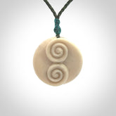 A hand carved and intricate koru pendant made for us by Yuri Terenyi. This is a beautiful little piece and is emblematic of the well known and loved Koru design. It is carved from bone in a round shape with decorative design carved into the koru. It is suspended from a sage green cord with a blue floret and the necklace is adjustable.