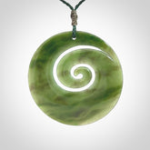 This picture shows a large hand-carved koru. It is carved from British Columbian jade and is suspended from a hand-plaited, green cord.