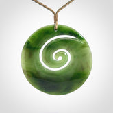 This picture shows a large hand-carved koru. It is carved from British Columbian jade and is suspended from a hand-plaited, tan/green cord.
