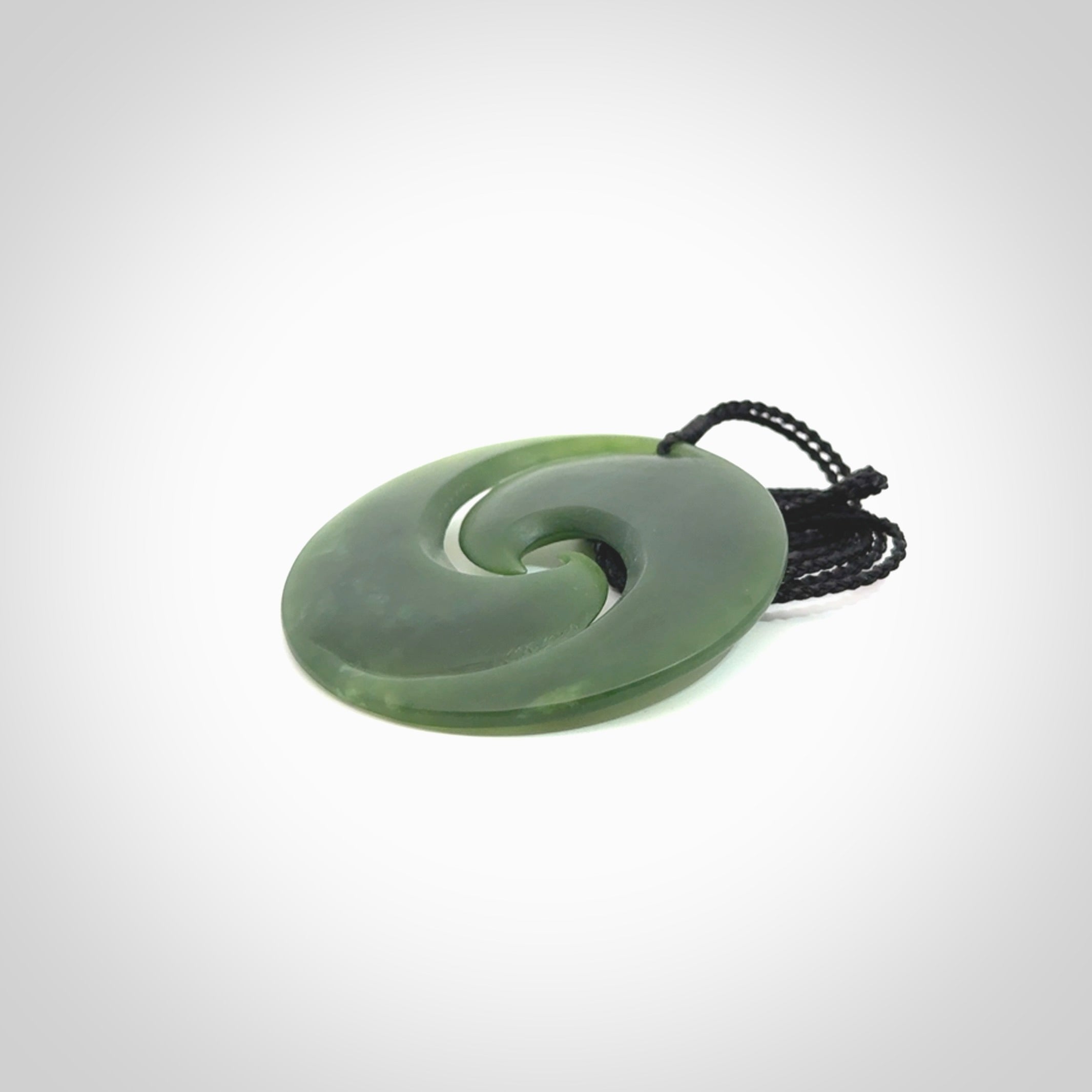 This picture shows a large hand carved jade koru pendant. It is a darker green colour which is quite translucent where the jade is thin. Although a large piece, it is a wonderful, delicate piece of jewellery. The cord is hand plaited and adjustable so that you can position the pendant where it suits you best.