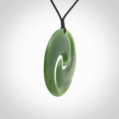This picture shows a large hand carved jade koru pendant. It is a darker green colour which is quite translucent where the jade is thin. Although a large piece, it is a wonderful, delicate piece of jewellery. The cord is hand plaited and adjustable so that you can position the pendant where it suits you best.