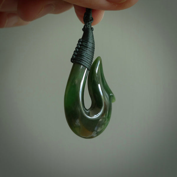 This photo shows a greenstone hook, or matau, pendant. It is a beautiful green British Columbia jade stone. The plaited cord is a manuka green colour. It is plaited and the length can be adjusted.