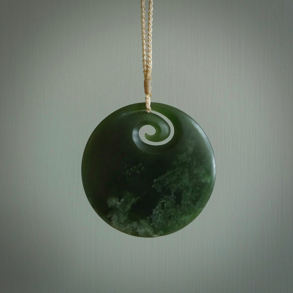 This picture shows a large hand carved jade koru pendant. It is a darker green colour which is quite translucent where the jade is thin. Although a large piece, it is a wonderful, delicate piece of jewellery. The cord is hand plaited and adjustable so that you can position the pendant where it suits you best. This koru was carved for us by Ric Moor. Delivery is free worldwide.
