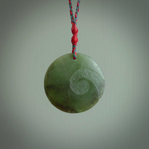 Hand carved jade disc pendant. One side has a single koru etched into the face. This is a subtle and beautiful design. The other side is plain. The cord is a Cobalt Blue/Juicy Red colour and is adjustable. The piece was carved for NZ Pacific by Rueben Tipene.