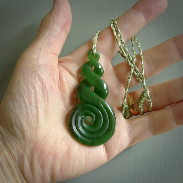 Ross Crump triple twist with koru pendant. Hand carved from rare New Zealand jade this is a beautiful pounamu pendant. The cord is hand plaited in green and white colours and is length adjustable. It has a floret in white. It is a delicate and very beautiful greenstone pendant. For sale online by NZ Pacific.