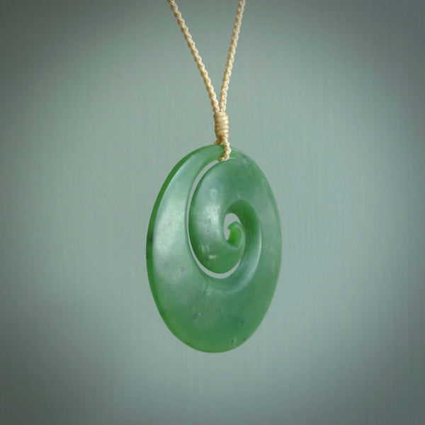 This picture shows a hand carved jade koru pendant. It is a deep green with orange colour and is a wonderful, large sized piece of jewellery. The cord is a four plait beige colour and adjustable so that you can position the pendant where it suits you best. This koru was carved for us by Ric Moor. Delivery is free worldwide.