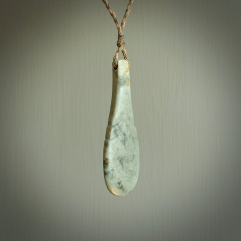 This is a sleek and slender hand carved New Zealand Jade mere pendant. The stone is a very dark green with mottled pale inclusions. The cord is tan and adjustable. The pendant has a light polish and just glows.