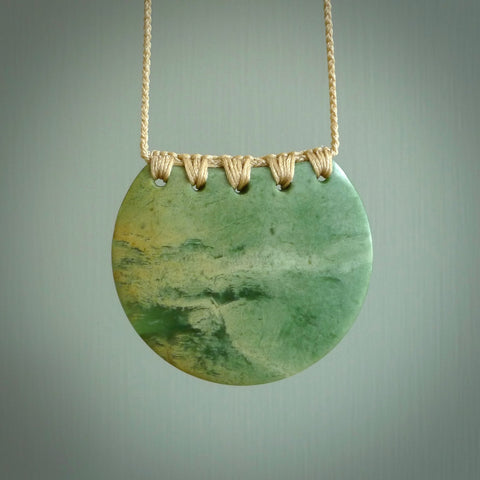 This piece is a large, oval round, disc pendant. It was carved for us by Ric Moor from a lovely milky green piece of New Zealand flower jade. It is suspended on a beige coloured braided cord that is length adjustable.