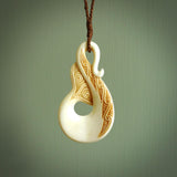 This picture shows a carved whale tail in bone. It is a hook shaped pendant pendant that culminates in a fish tail. The artist has carved traditional decorative Koru designs into the body and these run up the inner sides of the hook. These have specific meanings. It is provided with a plaited brown coloured cord that is length adjustable.