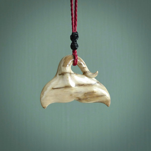This picture shows a hand carved whale tail pendant. It is a dark, ginger brown colour and is handcarved from Woolly Mammoth tusk. The necklace is made from a braided cord in a pohutukawa red colour.