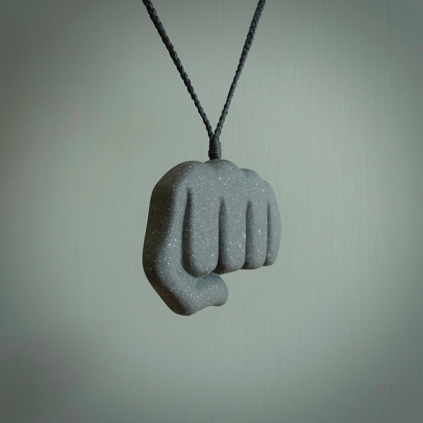 Greywacke Beach Stone fist bump, pendant hand made here in New Zealand. Hand carved by Rhys Hall for NZ Pacific. Handmade jewellery for sale online. The cord is black and has a loop and pebble toggle closure. Unique necklace for men and women. fist pump necklace hand made from New Zealand Greywacke Beach Stone.