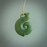 A hand carved hei-matau with koru pendant. This pendant is a green and orange colour and is finished in a polished matte. The cord is a four plait in beige and it is adjustable.