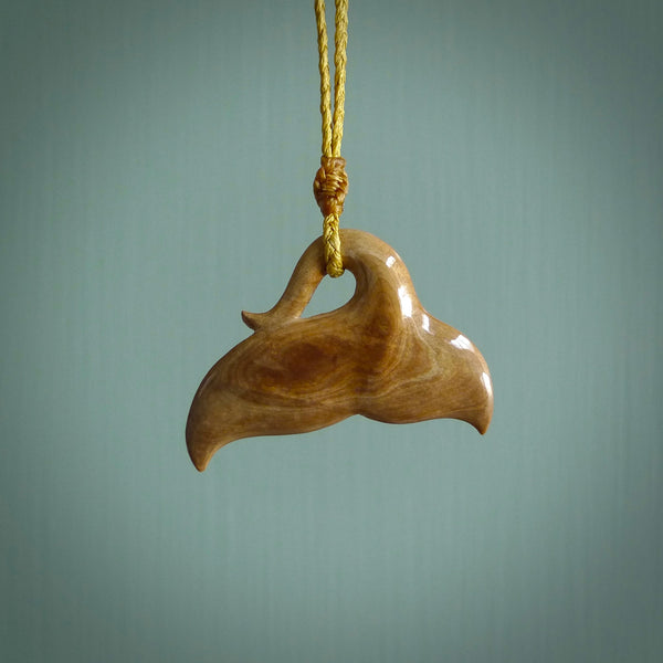 This picture shows a hand carved whale tail pendant. It is a dark, ginger brown colour and is handcarved from Woolly Mammoth tusk. The necklace is made from a braided cord in a mustard colour.