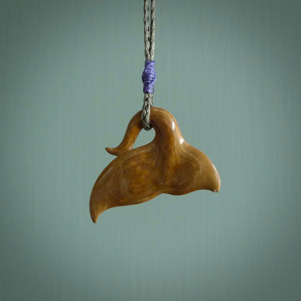 This picture shows a hand carved whale tail pendant. It is a dark, ginger brown colour and is handcarved from Woolly Mammoth tusk. The necklace is made from a braided cord in an olive green colour.