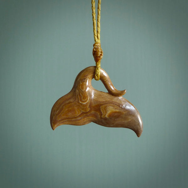 This picture shows a hand carved whale tail pendant. It is a dark, ginger brown colour and is handcarved from Woolly Mammoth tusk. The necklace is made from a braided cord in a mustard colour.