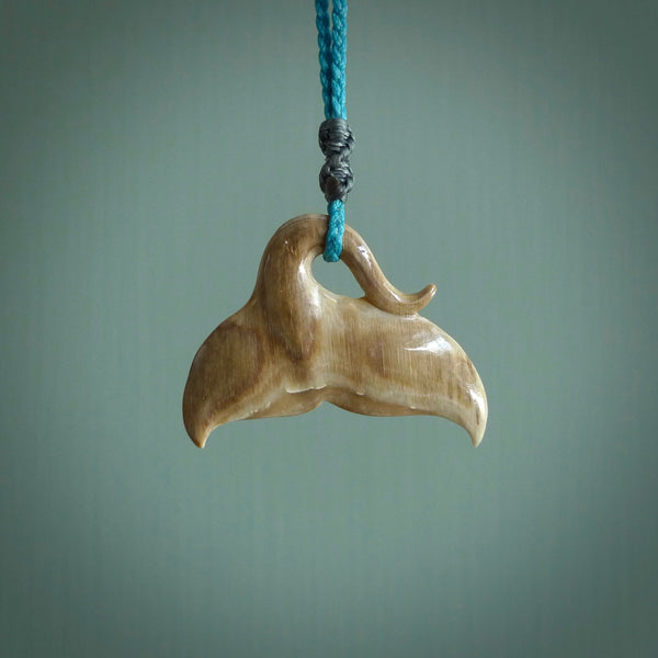 This picture shows a hand carved whale tail pendant. It is a dark, ginger brown colour and is handcarved from Woolly Mammoth tusk. The necklace is made from a braided cord in a paradise blue colour.