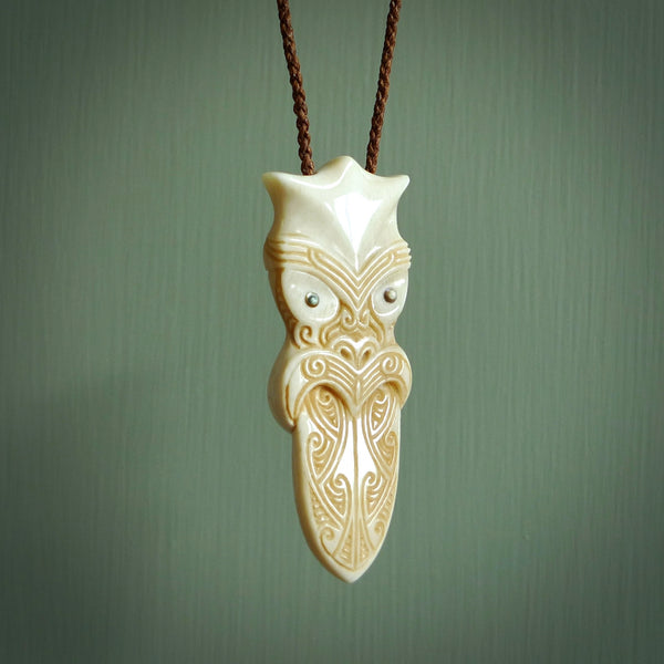 This is a large carved Koruru pendant. It is made from two materials - New Zealand Paua Shell and Bone. The face of the Koruru is carved with a traditional design in the front. It has a protruding tongue which is also carved and the back face has traditional carved designs. This is a large and very traditional pendant that is a collectors piece. This piece is provided on a brown adjustable cord.