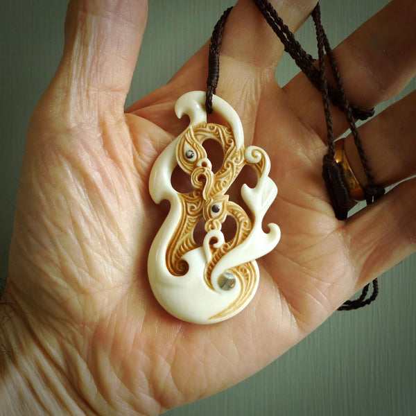 This picture shows a carved double manaia in bone. The artist has carved traditional decorative Koru designs into the face of the body and these run up the sides of the manaia head. These have specific meanings. It is provided with a hand-plaited brown cord that is length adjustable.