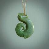 A hand carved hei-matau with koru pendant. This pendant is a green and orange colour and is finished in a polished matte. The cord is a four plait in beige and it is adjustable.