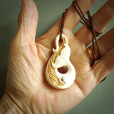 This picture shows a carved whale tail in bone. It is a hook shaped pendant pendant that culminates in a fish tail. The artist has carved traditional decorative Koru designs into the body and these run up the inner sides of the hook. These have specific meanings. It is provided with a plaited brown coloured cord that is length adjustable.