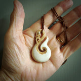 This picture shows a carved manaia in bone. It is a hook shaped pendant pendant that culminates in a fish tail. The artist has carved traditional decorative Koru designs into the side of the body and these run up the sides of the manaia head. These have specific meanings. It is provided with a hand-plaited brown cord that is length adjustable.