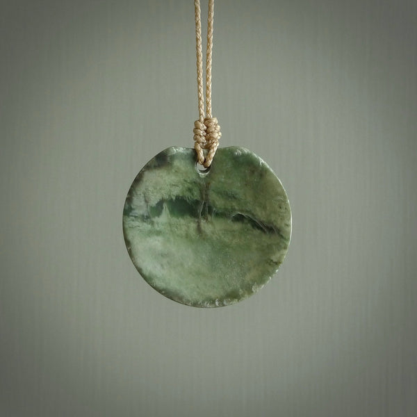 This piece is a oval round, disc pendant. It was carved for us by Ric Moor from a lovely light and milky green piece of New Zealand jade. It is suspended on a beige coloured braided cord that is length adjustable.