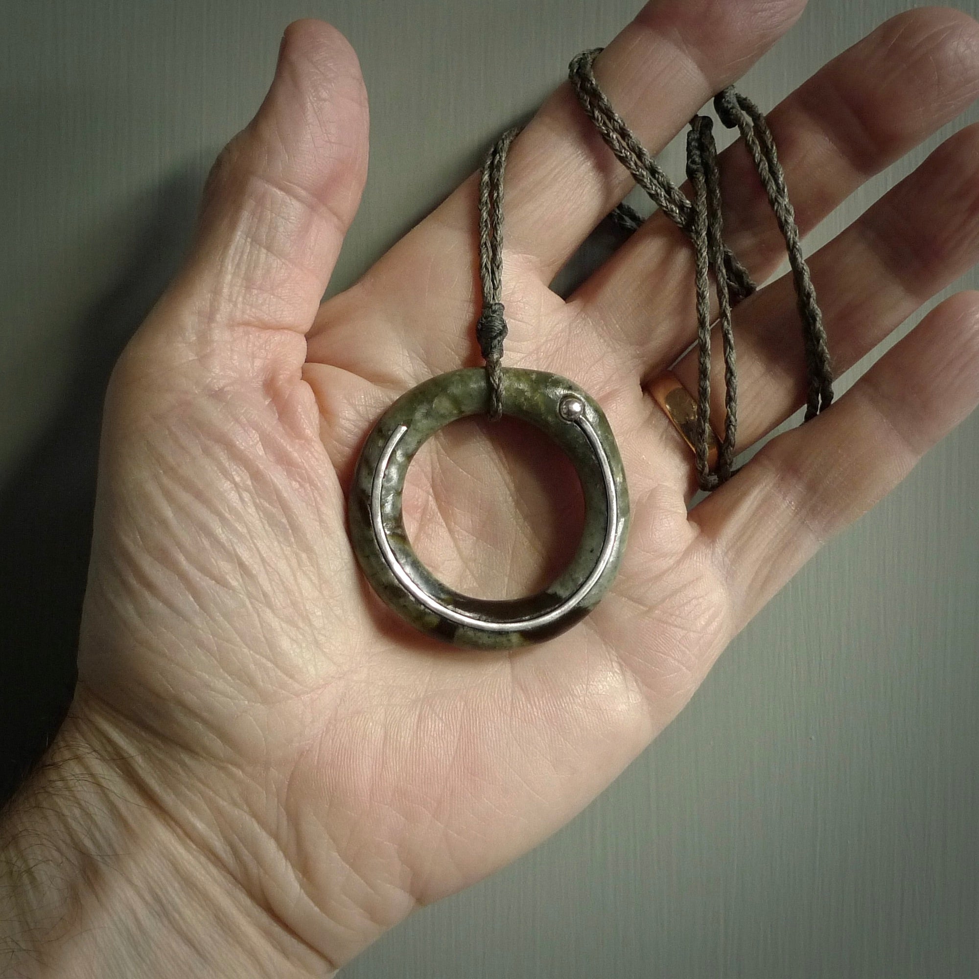This picture shows a hand carved jade circle pendant with sterling silver inlay. It is suspended from an adjustable Manuka Green and Tan coloured cord. Delivery is free worldwide.