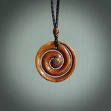 A hand carved and intricate koru pendant made for us by Yuri Terenyi. This is a beautiful little piece and is emblematic of the well known and loved Koru design. It is carved from stained bone in a hollowed, koru spiral shape with paua shell inlay in the middle of the koru. It is suspended from a black cord with a gingernut floret and the necklace is adjustable.