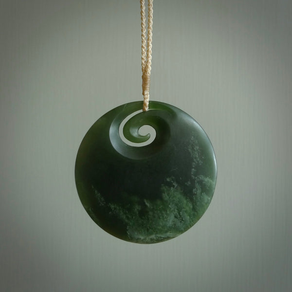 This picture shows a large hand carved jade koru pendant. It is a darker green colour which is quite translucent where the jade is thin. Although a large piece, it is a wonderful, delicate piece of jewellery. The cord is hand plaited and adjustable so that you can position the pendant where it suits you best. This koru was carved for us by Ric Moor. Delivery is free worldwide.