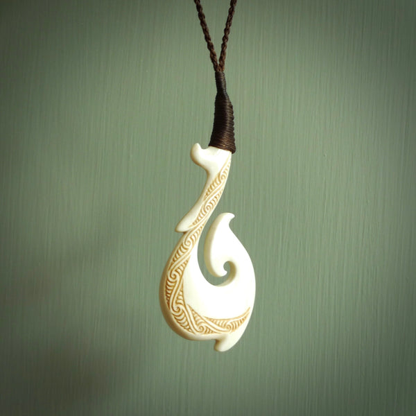 This picture shows a carved hook in bone. The artist has carved traditional decorative kowhaiwhai designs into the body and these run up the insides of the matau. These have specific meanings. It is provided with a hand-plaited brown cord that is length adjustable. Free shipping worldwide.