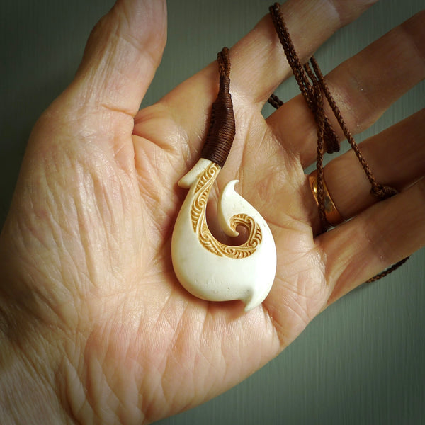 This picture shows a carved hook in bone. The artist has carved traditional decorative kowhaiwhai designs into the body and these run up the insides of the matau. These have specific meanings. It is provided with a hand-plaited brown cord that is length adjustable. Free shipping worldwide.