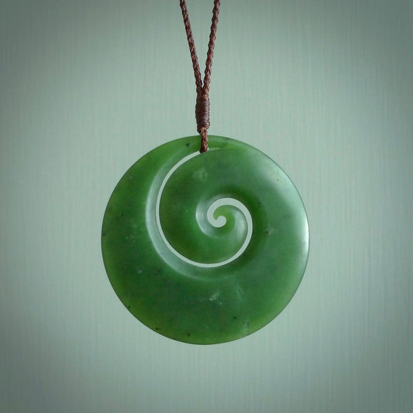 This picture shows a large hand carved jade koru pendant. It is a darker green colour which is quite translucent where the jade is thin. Although a large piece, it is a wonderful, delicate piece of jewellery. The cord is hand plaited and adjustable so that you can position the pendant where it suits you best. This koru was carved for us by Ric Moor. Delivery is free worldwide.
