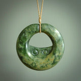 This piece is a large sized, oval round, disc pendant with double koru heart. It was carved for us by Ric Moor from a lovely deep and milky green piece of New Zealand flower jade. It is suspended on a beige  coloured braided cord that is length adjustable.
