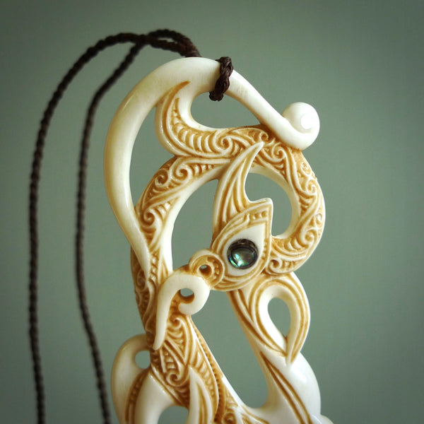 This picture shows a carved double manaia in bone. The artist has carved traditional decorative Koru designs into the face of the body and these run up the sides of the manaia head. These have specific meanings. It is provided with a hand-plaited brown cord that is length adjustable.