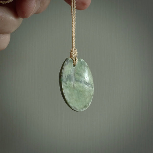 This piece is a oval round, disc pendant. It was carved for us by Ric Moor from a lovely light and milky green piece of New Zealand jade. It is suspended on a beige coloured braided cord that is length adjustable.