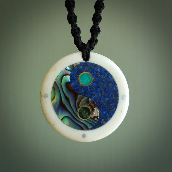 This photo shows a medium sized yin and yang pendant hand carved from bone with New Zealand Pounamu Jade, Lapis Lazuli and Paua Shell inlay alongside; brass and silver. This is a stand out one off necklace for those who appreciate art to wear. It is provided with a cord in black that is a fixed length with Paua Shell Toggle. We ship this piece worldwide and shipping is included in the price.