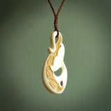 This picture shows a carved whale tail in bone. It is a hook shaped pendant pendant that culminates in a fish tail. The artist has carved traditional decorative Koru designs into the body and these run up the inner sides of the hook. These have specific meanings. It is provided with a plaited brown coloured cord that is length adjustable.