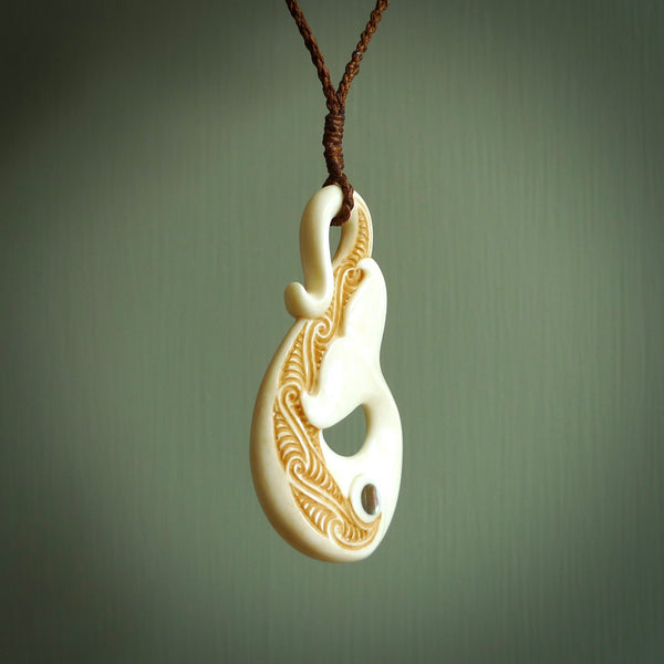 This picture shows a carved whale tail in bone. It is a hook shaped pendant pendant that culminates in a fish tail. The artist has carved traditional decorative Koru designs into the body and these run up the inner sides of the hook. These have specific meanings. It is provided with a plaited brown coloured cord that is length adjustable.