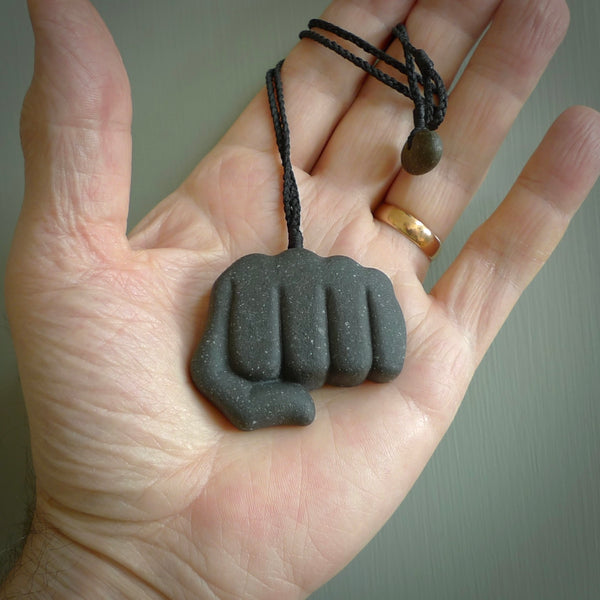 Greywacke Beach Stone fist bump, pendant hand made here in New Zealand. Hand carved by Rhys Hall for NZ Pacific. Handmade jewellery for sale online. The cord is black and has a loop and pebble toggle closure. Unique necklace for men and women. fist pump necklace hand made from New Zealand Greywacke Beach Stone.